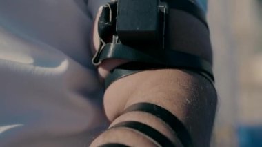 An elderly Jewish man is sitting on the chair outside with a tefillin on his hand.