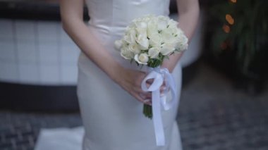 Elegant bride in a white dress holding a beautiful wedding bouquet of fresh white roses. Romantic bridal details, wedding ceremony preparation, love and celebration concept.