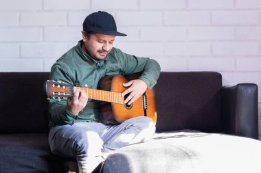 Kanepede oturan adam sol eliyle gitar çalıyor.
