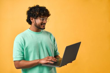 Turkuaz tişörtlü ve gözlüklü yakışıklı bir Hintli adamın portresi bir dizüstü bilgisayar tutuyor ve sarı bir arkaplanda dururken daktilo çekiyor..                               