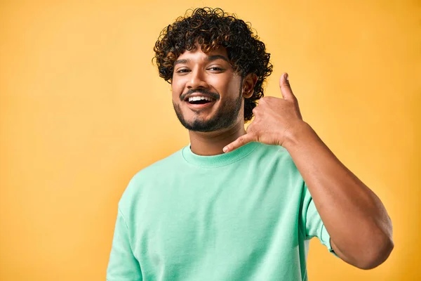 A candid portrait of a handsome curly-haired young man in a turquoise t-shirt and on a yellow background who is laughing and making a hand gesture to call him.