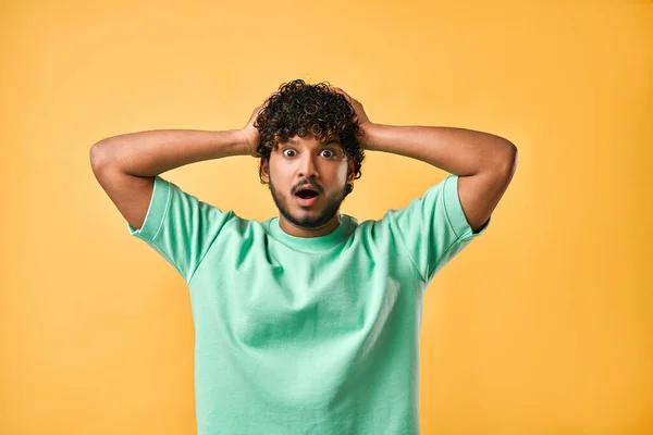 stock image Portrait of a handsome young man in a turquoise T-shirt on a yellow background holding his head and expressing the emotion of surprise, fear, shock.