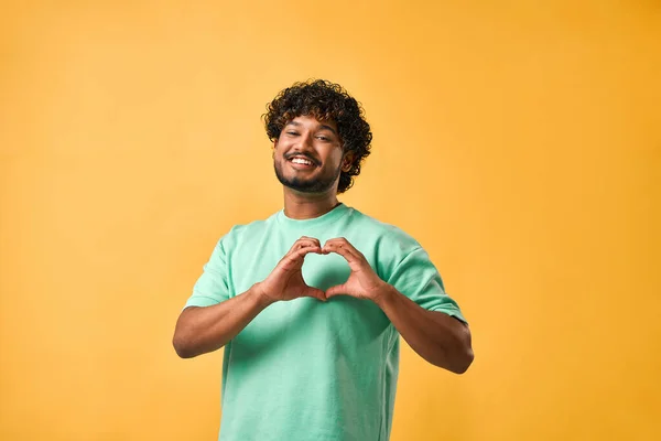 An image of an Indian man in a turquoise t-shirt showing a gesture of heart, love, affection, care with his fingers and smiling while looking at the camera.