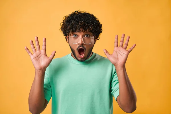 stock image Close-up portrait of handsome Indian man in turquoise t-shirt and glasses with big eyes and open mouth standing on yellow background and expressing emotion of shock, surprise and showing hands.