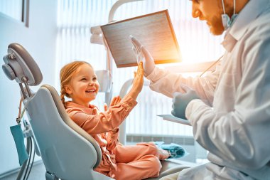A little girl is sitting in a dentist's chair, giving a high five to the doctor and laughing. Dental care, trust and patient care. Children's dentistry.Sunlight.                                clipart