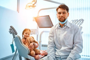 Diş muayenehanesinde oturmuş gülümseyen bir dişçinin portresi. Arka planda elinde oyuncak tutan küçük bir kız hasta. Çocuk dişçiliği. Güneş ışığı..