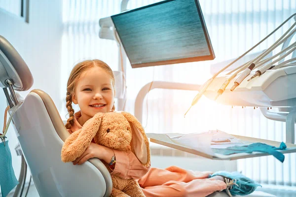 stock image A cute little girl sitting in a dental chair, holding a toy rabbit and laughing. In the background, dental equipment and a monitor for the clinic logo. Dental care, medicine.Sunlight.