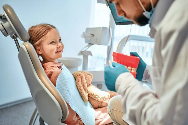 Stock image Children's dentistry. A little girl sits in a dental chair and looks at the doctor who shows her a model of the jaw and shows how to brush her teeth.