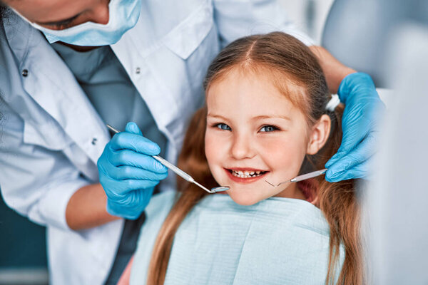 Photo of a child smiling at a dentist appointment. A doctor in gloves holds examination tools behind. Children's dentistry.                               