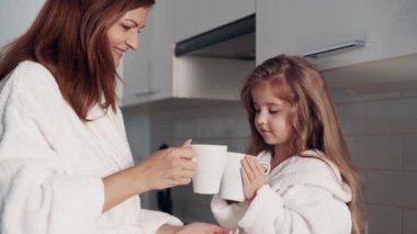 Genç bir anne ve anaokulu kızı mutfaktaki kupalardan çay içiyorlar..