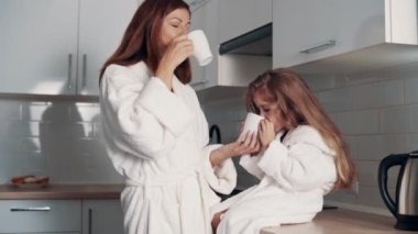 Genç bir anne ve anaokulu kızı mutfaktaki kupalardan çay içiyorlar..