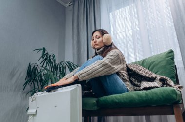 Sıcak giyimli genç bir dişi evdeki elektrikli radyatörün yanında ısınıyor..