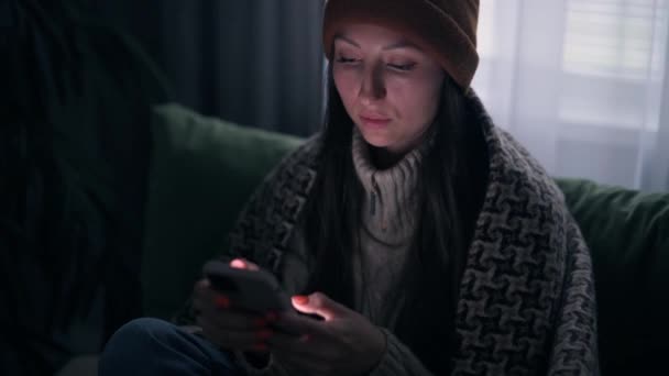 暖かい服装の若い女性は電気や暖房のない暗い部屋でスマートフォンを使用しています 緊急停止 — ストック動画