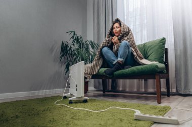 Sıcak giyimli genç bir dişi evdeki elektrikli radyatörün yanında ısınıyor..