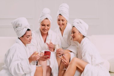 A group of women spend leisure time. Four Young attractive women in bathrobes look at the smartphone screen and smile.