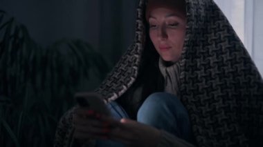 Sıcak giyimli genç bir kadın karanlık bir odada elektrik ve ısıtma olmadan akıllı telefon kullanıyor. Acil kapatma.