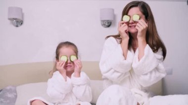 Anne ve kızı evde dinleniyor. Genç bir kadın ve dört yaşındaki kızı göz kapaklarında salatalık yüzükleriyle bir yatakta oturuyorlar..