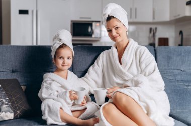 Genç bir anne ve anaokulu kızı evdeki kanepede fincanlardan çay içiyorlar..