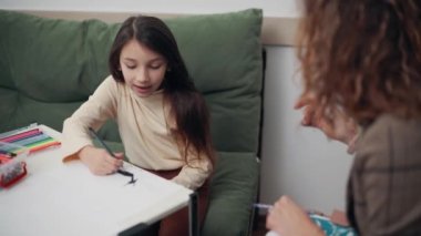 Woman Psychologist communicates with a girl in the office. The child draws and talks with the doctor.