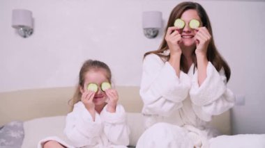 Anne ve kızı evde dinleniyor. Genç bir kadın ve dört yaşındaki kızı göz kapaklarında salatalık yüzükleriyle bir yatakta oturuyorlar..