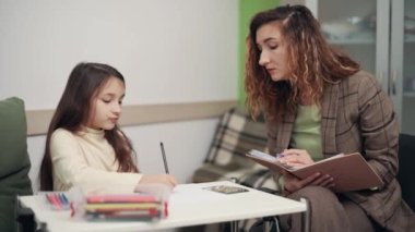 Woman Psychologist communicates with a girl in the office. The child draws and talks with the doctor.