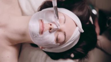 Kozmetik uzmanı müşterilerin yüzüne nemlendirici bir maske takıyor. Genç bir kadın kaplıcada rahat bir şekilde uzanıyor. Üst görünümü kapat.