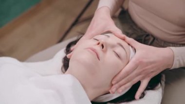 Güzellik uzmanı yüz masajı yapıyor. Genç bir kadın sırt üstü uzanıyor ve kaplıca salonunda yüzünü gerdiriyor. Yaşlanmaya karşı masaj teknikleri.