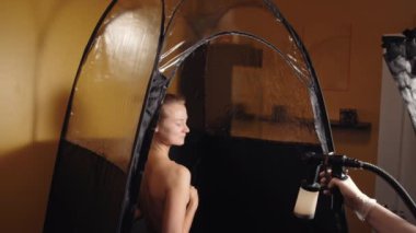 Cosmetologist using airbrush for spray tan apply to young woman body in beauty salon. 