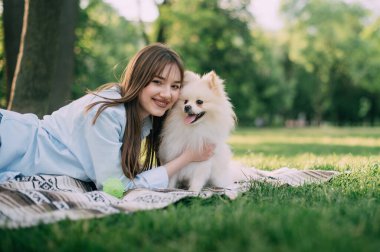 Genç bir kadın çimenlerde uzanıyor ve Pomeranya köpeğini kollarında tutuyor..