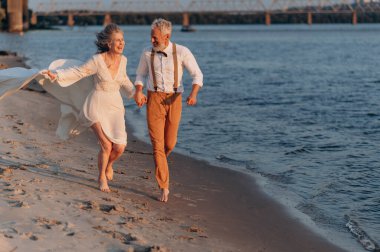 Neşeli yaşlı çift kumsalda el ele koşuyor..