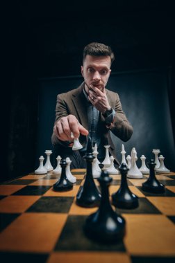 Konsantre sakallı adam satrançta hamle yapıyor. Siyah arkaplanda dramatik bir portre.