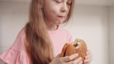 Küçük bir kız mutfakta iştahlı büyük bir hamburger yer. Kapat..