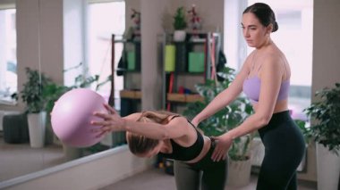 Pilates yapan kadınlar. Antrenör, müşterinin jimnastik topuyla egzersiz yapmasına yardım eder..