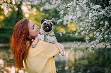 Genç kızıl saçlı bir kadın köpeğini önünde tutuyor. Sahibi ve evcil hayvanı dışarıda eğleniyorlar.. 