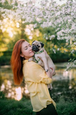 Genç kızıl saçlı bir kadın köpeğini önünde tutuyor. Sahibi ve evcil hayvanı dışarıda eğleniyorlar.. 
