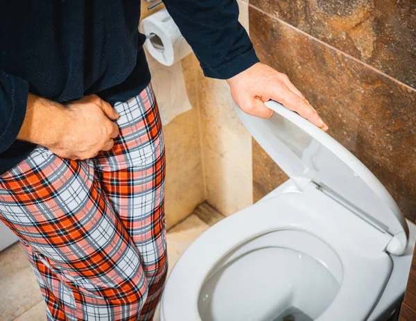 A man in pajamas in the toilet squeezes his crotch and lifts the toilet seat, Urination problem,  Health concept, Male prostate problem, urinary system function, bladder pain