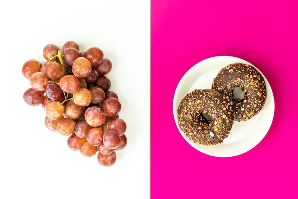 Healthy and unhealthy food, flat layer, two color background, chocolate donuts with sprinkles on a fuchsia background, juicy delicious grapes on white, Natural sugar vs harmful processed