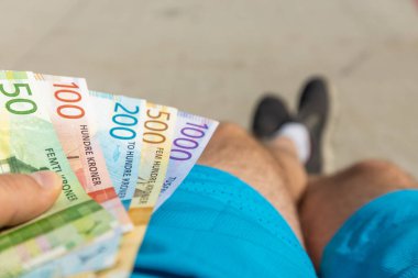 Bir bankta oturan genç bir adam elinde Norveç, Norveç, finansal konsept, ilk iş, finansal sorumluluk gibi bir tomar Norveç kronunu tutuyor.