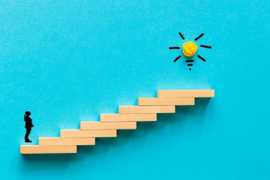 Woman standing in front of stairs leading up with a Glowing Light Bulb icon at the top. Achieving goals and objectives or goal setting concept. Blue background, copy space