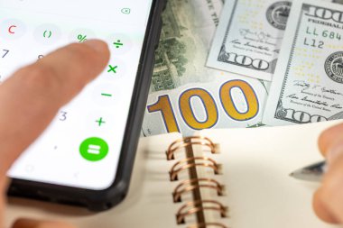 Amerikan doları. Akıllı telefon hesap makinesine güvenerek, elinde kalem ve defterle. Amerikan doları. Finansal yerleşim, Ev bütçesi, Vergiler, ABD döviz kuru, İş Analizi Konsepti