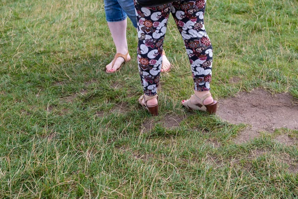 two women stand on a meadow, one with high wedge heels shoes and summer trousers and the other with flip-flops and wise shorts.