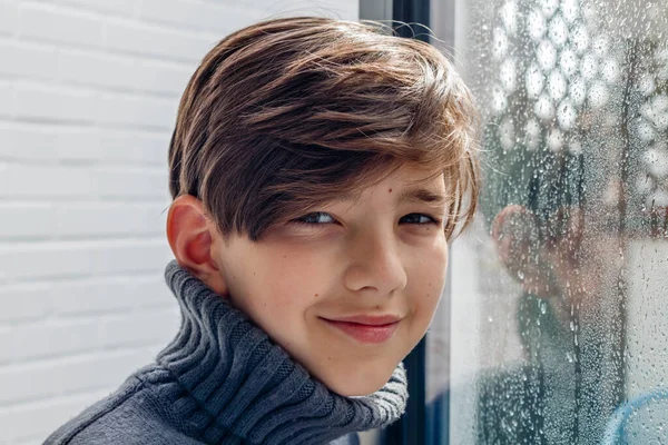 Smiling boy in grey turtleneck sweater standing by the window with rainy drops and looking at camera. Reflection