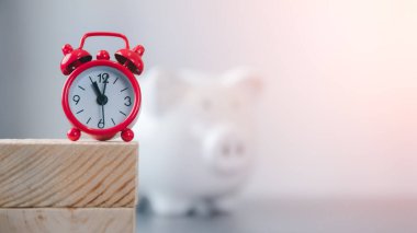 A red clock rests on a stick and piggy banks both rest on the table. Financial and money deposit concept.