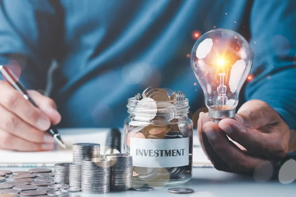 light bulb in a businessman\'s hand and a pile of coins and coins in a glass jar.All on table,money saving and investment ideas and profit growth,financial management and business strategy.for success