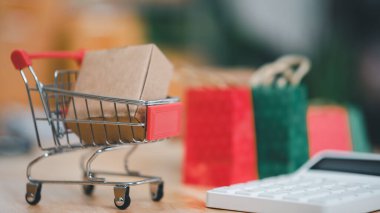 Alışveriş arabası, karton kutu ve hesap makinesi masanın üzerinde, Tüketici Derneği, İnternet üzerinden alışveriş hizmeti ve ev teslimatı, çevrimiçi alışveriş veya e-alışveriş sunar