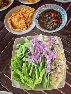 Bir tabak taze yeşil sebze ve mor çiçekten oluşan canlı bir yemek, sulu bir omlet ve kırmızı biberli baharatlı bir sos tabağı ve karabiber..