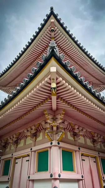 Etkileyici bir geleneksel Japon mimarisi yapısı, özenle hazırlanmış katmanlı çatılar, karmaşık pembe ve yeşil detaylarla. Görüntü binanın simetrik güzelliğini yakalıyor ve bulutlu hava manzaraya dramatik bir dokunuş ekliyor.