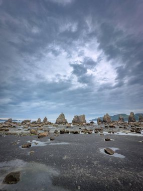 Bulutlu bir gökyüzünün altındaki kayalık bir sahilin panoramik görüntüsü. Kum üzerinde yükselen kaya oluşumları, doğanın sükunetini ve güzelliğini yansıtıyor, dinginlik ve gizemi çağrıştırıyor..