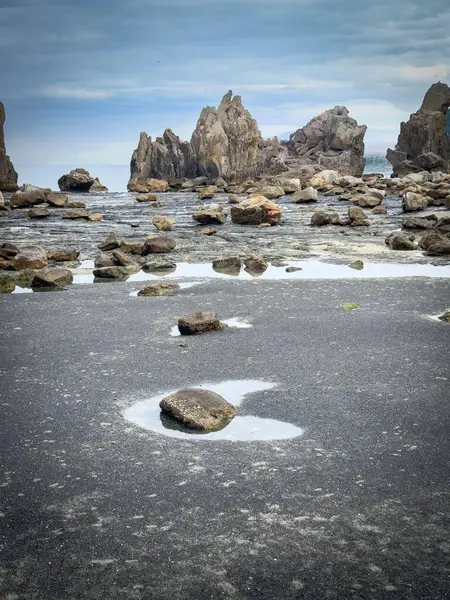 Arkaplanda su ve gök mavisi renkleriyle çevrili, bulutlu bir gökyüzünü yansıtan büyük taş oluşumları olan kayalık bir sahilin panoramik görüntüsü. Sahne huzuru ve doğanın gücünü çağrıştırıyor..