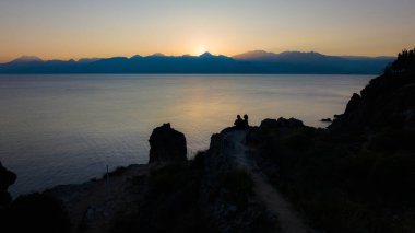 Gün batımında romantik çift silueti. Dağların arasında batan kızıl güneş. Deniz kenarında